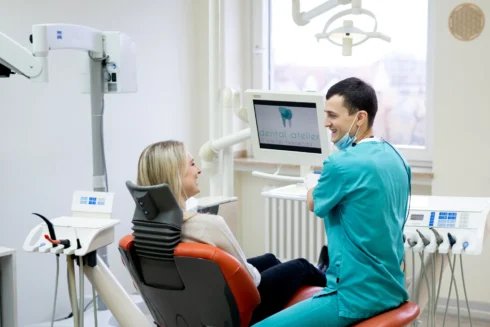 Junge, blonde Patientin  sitzt auf Behandlungsstuhl neben ihr sitzt Dmitri Tanurcov und zeigt ihr etwas auf einem Display am Stuhl