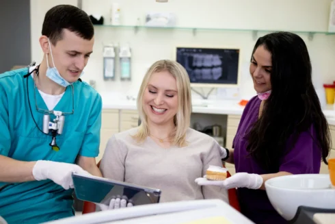 Blonde, lächelnde Patientin sitzt auf Behandlungsstuhl. Dmitri Tanurcov zeigt ihr Unterlagen. Daneben steh eine ZFA mit einem Zahnmodell in der Hand